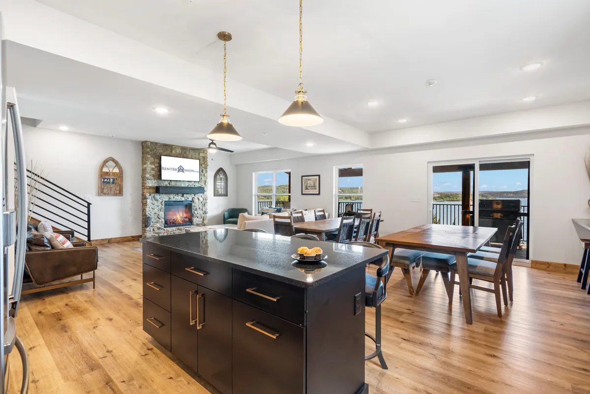 An expansive open-concept area blending the kitchen, dining, and living room. A central kitchen island overlooks a spacious dining area, a cozy fireplace, and large windows letting in ample natural light. The scene exudes warmth and sociability, with comfortable seating arranged for conversation or relaxation.