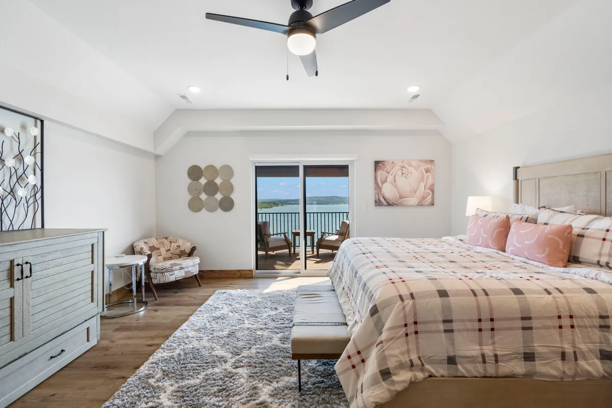 A serene king-sized bedroom featuring a large bed with plush bedding, a sliding glass door that opens to scenic views, tasteful decorative artwork, and a cozy nightstand. The room is filled with natural light, suggesting a restful and inviting atmosphere.