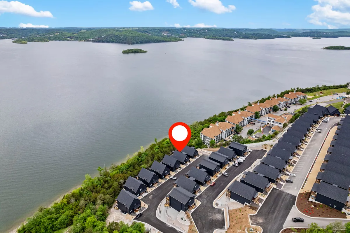 A peaceful lakeshore scene with a wide view of the lake in the foreground, bordered by a cluster of modern townhouses and lush green landscaping. The sky is calm, and the water reflects the serenity of the surrounding environment. This location is ideal for boating, fishing, or simply relaxing by the water with family and friends.