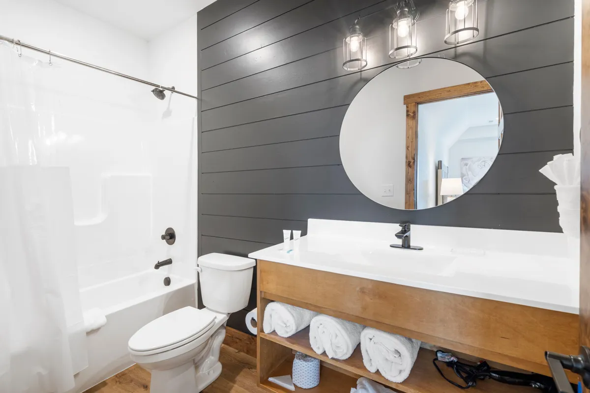 A modern bathroom featuring a round mirror, wooden vanity, and a visible walk-in shower with accessible design. The space is bright and fresh, reflecting a commitment to comfort and inclusivity for all guests, including those with mobility needs.