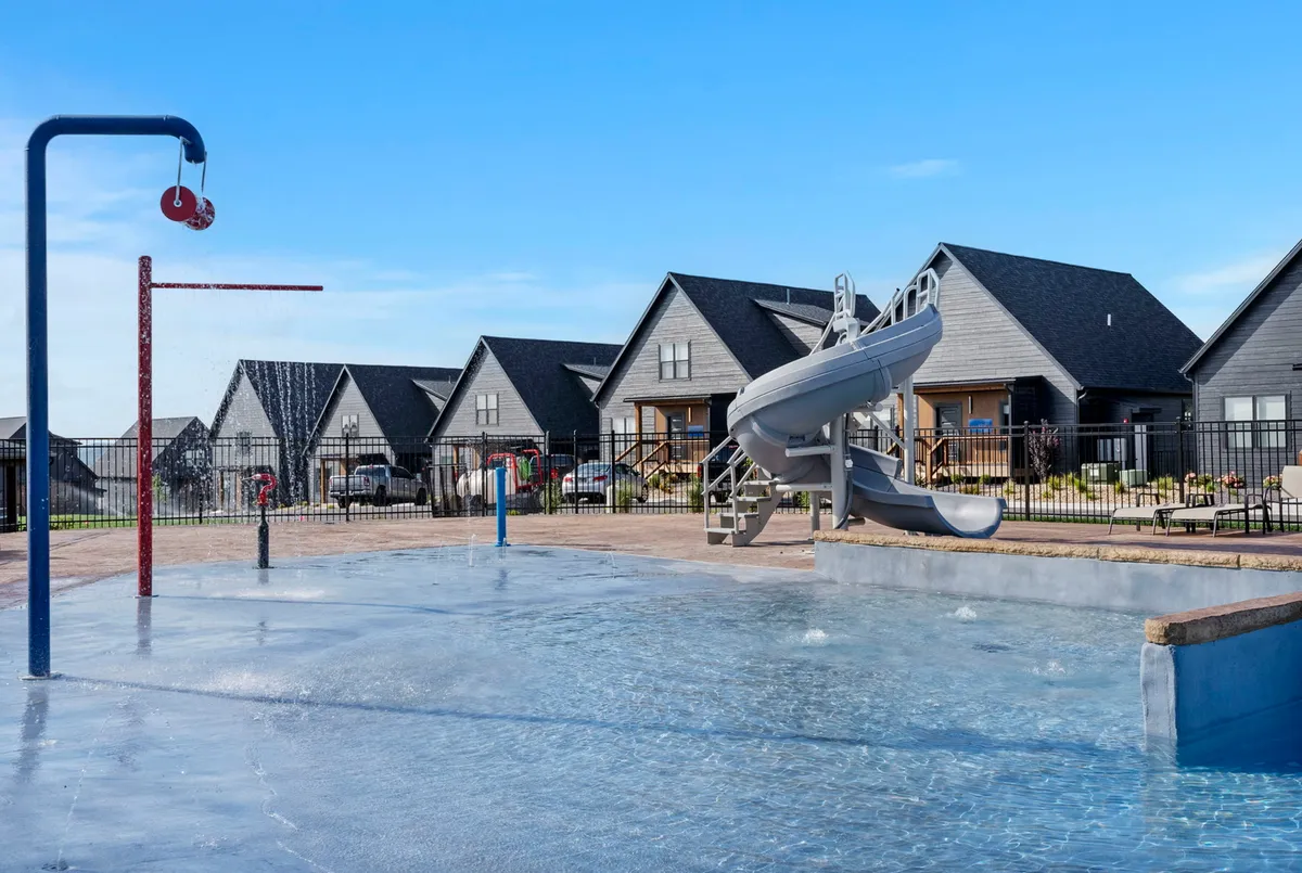 A vibrant outdoor swimming pool area features a colorful water slide, spray features, and plenty of lounge chairs. Children are seen playing in the water while families gather around the pool. The surrounding houses create a communal atmosphere perfect for group activities and summer fun.