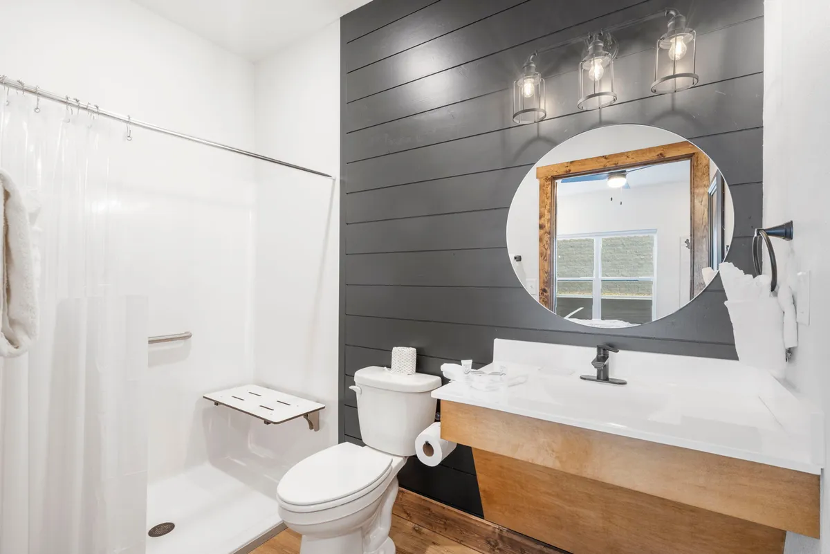 A contemporary bathroom with a circular mirrored sink, wooden vanity, black shiplap accent wall, and stylish metal light fixtures. The space is clean, modern, and inviting—ideal for refreshing after a day by the lake.